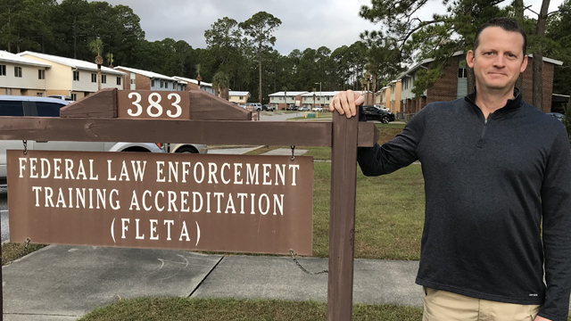 JJ Hensley stands next to the Federal Law Enforcement Training Accreditation Offic of Accreditation sign